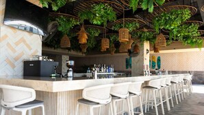 Un espace bar doté d’un long comptoir, de tabourets de bar blancs, de luminaires suspendus en osier et d’un mur orné d’un motif en chevrons.