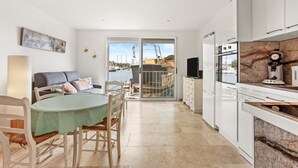 A modern kitchen with white cabinets, a dining area with a green tablecloth, and a view of boats through glass doors. - Very pleasant ground-floor apartment near the beach (Grimaud)