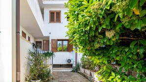 Un patio con plantas en macetas, un edificio blanco con contraventanas marrones y un equipo de aire acondicionado.