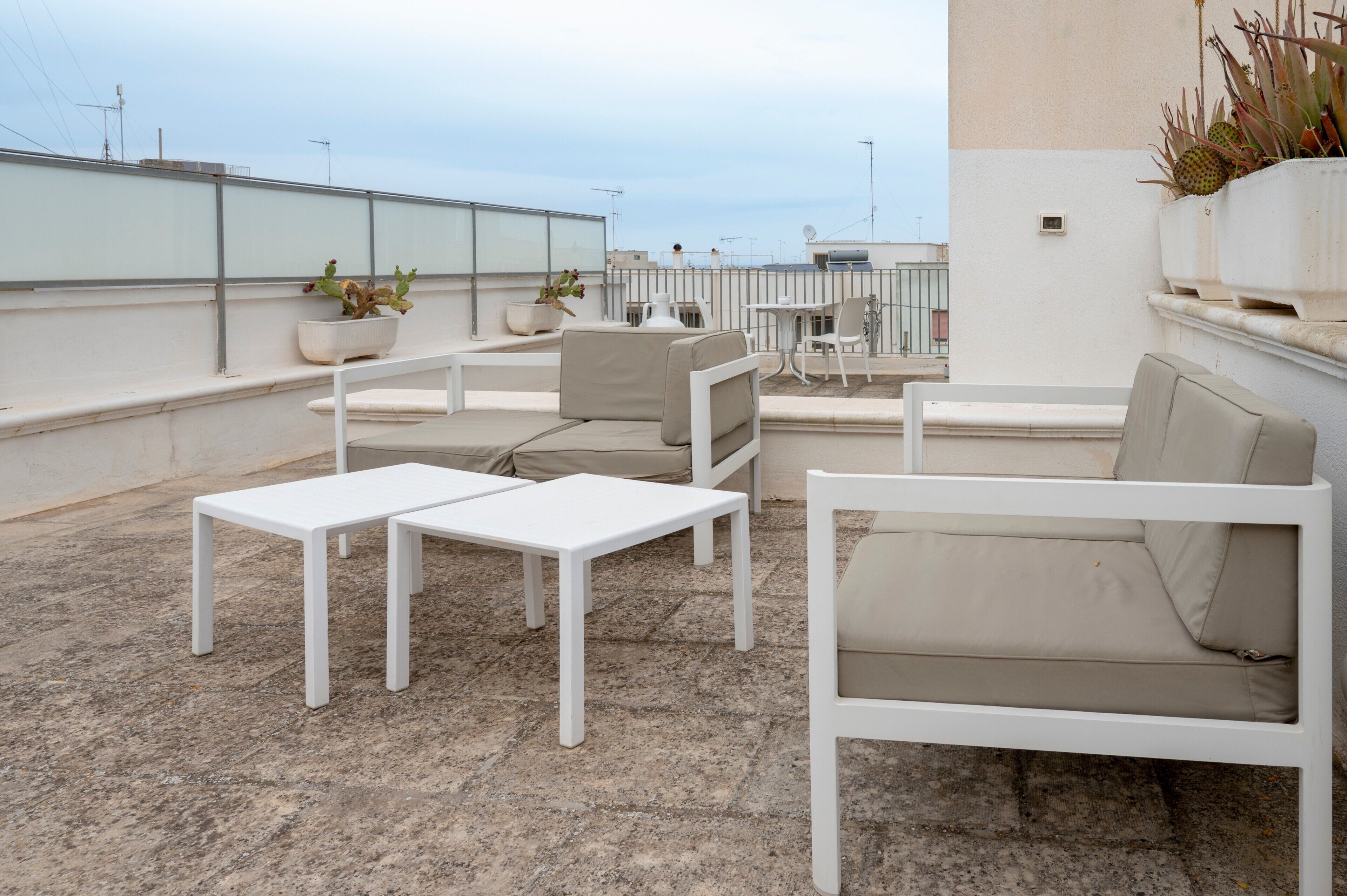 Terraza en la azotea con mobiliario blanco, que incluye un sofá y dos mesas, y plantas en macetas.
