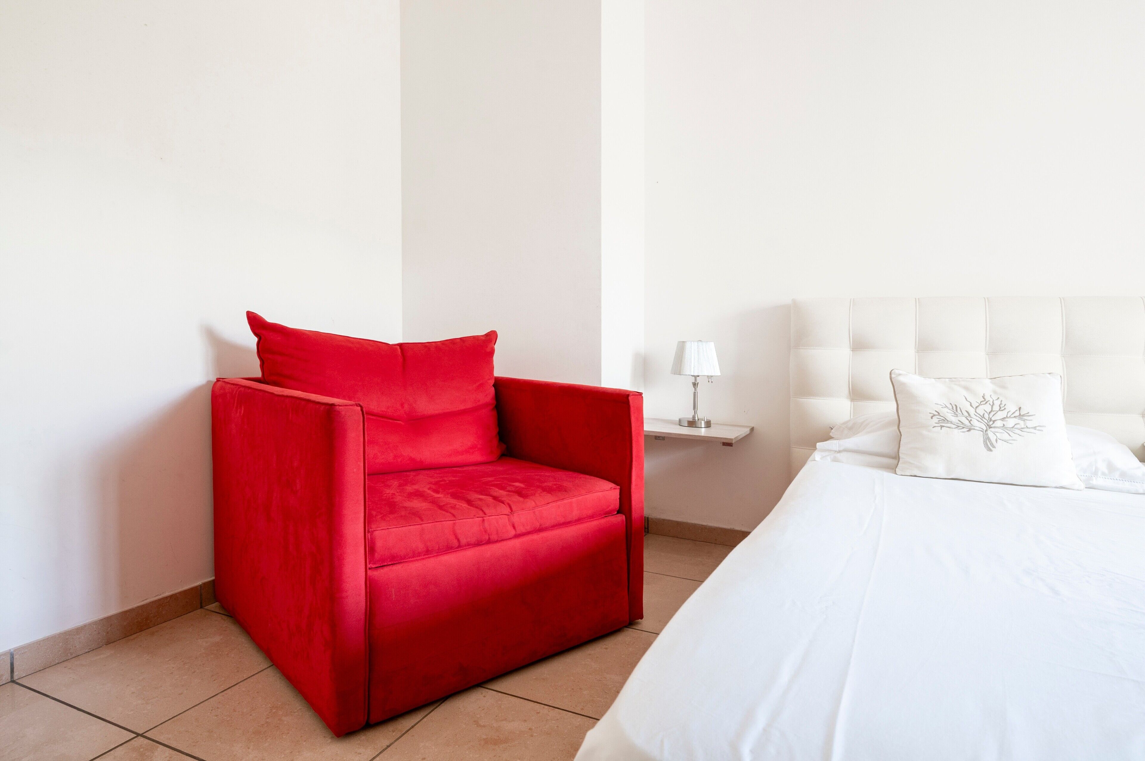 Une chambre d’hôtel avec un fauteuil rouge, un lit blanc et une table de chevet avec une lampe.