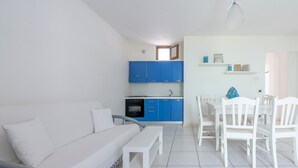 Un saló modern amb un sofà blanc, una taula de menjador amb caires blanques i una cuina amb armaris blaus.