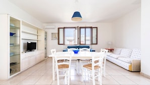 Une cuisine moderne avec des meubles blancs, une table à manger avec des chaises, un luminaire suspendu bleu et un canapé à motifs.