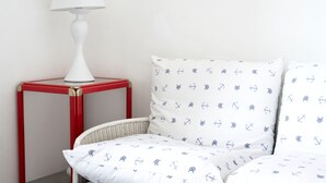 Une chaise en osier blanche avec un coussin blanc orné d’un motif nautique, une lampe de table blanche posée sur une table rouge et un mur blanc uni.