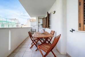 Een balkon met een houten tafel en stoelen, een witte muur en een raam met houten luiken.