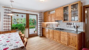 Une cuisine avec des meubles en bois, une table à manger recouverte d’une nappe à motifs et une vue sur la verdure à l’extérieur.