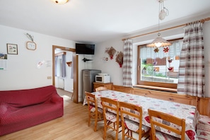 A cozy living room with a red sofa, wooden dining table with chairs, a TV, and a window with curtains.
