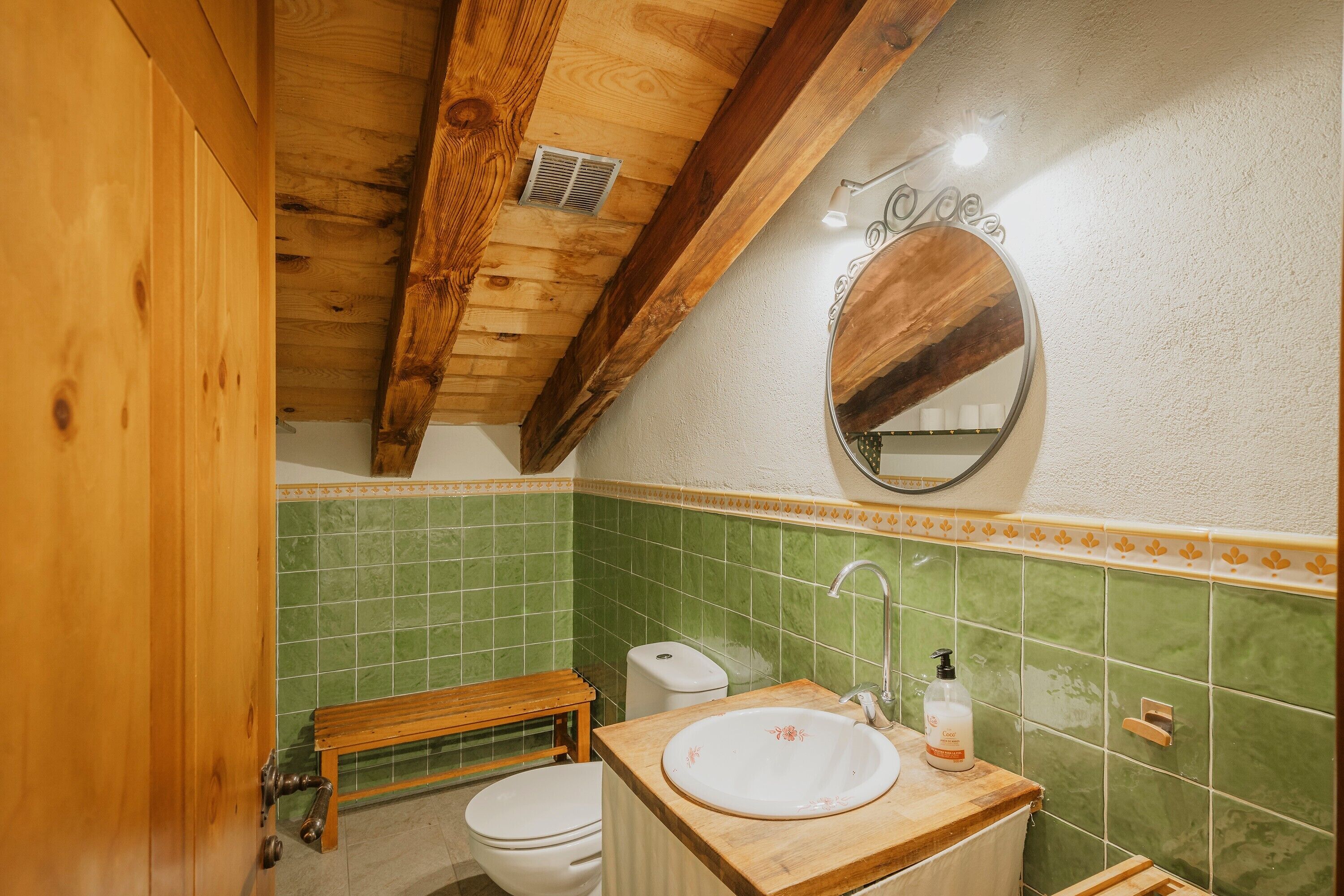 A bathroom with a wooden ceiling, green tiled walls, a wooden bench, a toilet, and a sink with a mirror above it.