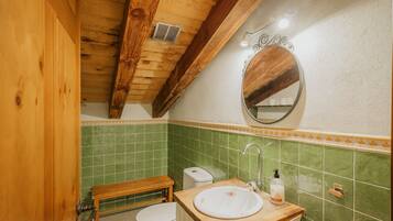 A bathroom with a wooden ceiling, green tiled walls, a wooden bench, a toilet, and a sink with a mirror above it.