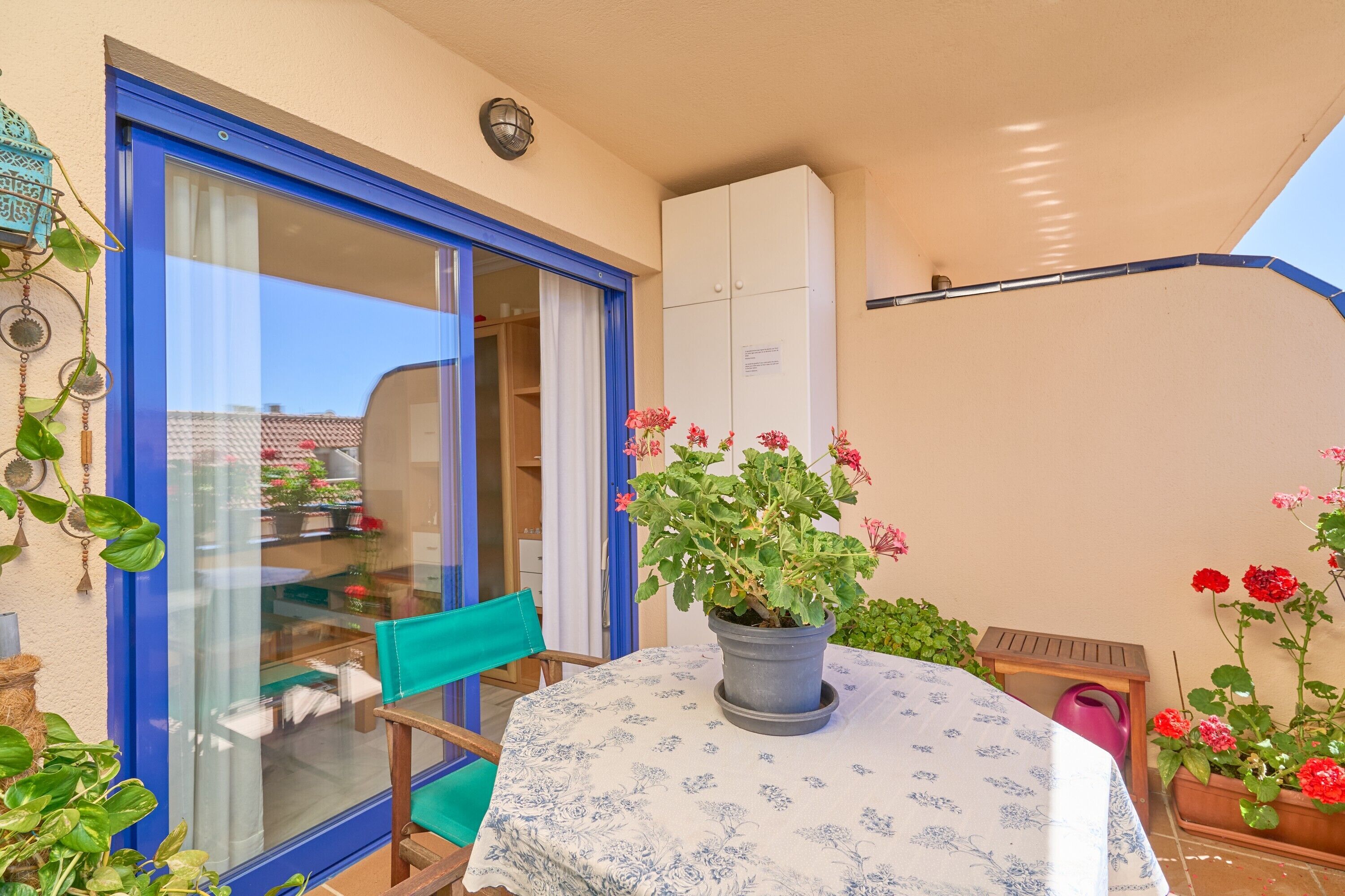 Un pequeño patio con una mesa, sillas, una planta en maceta y un ventanal corredero que da a una habitación.
