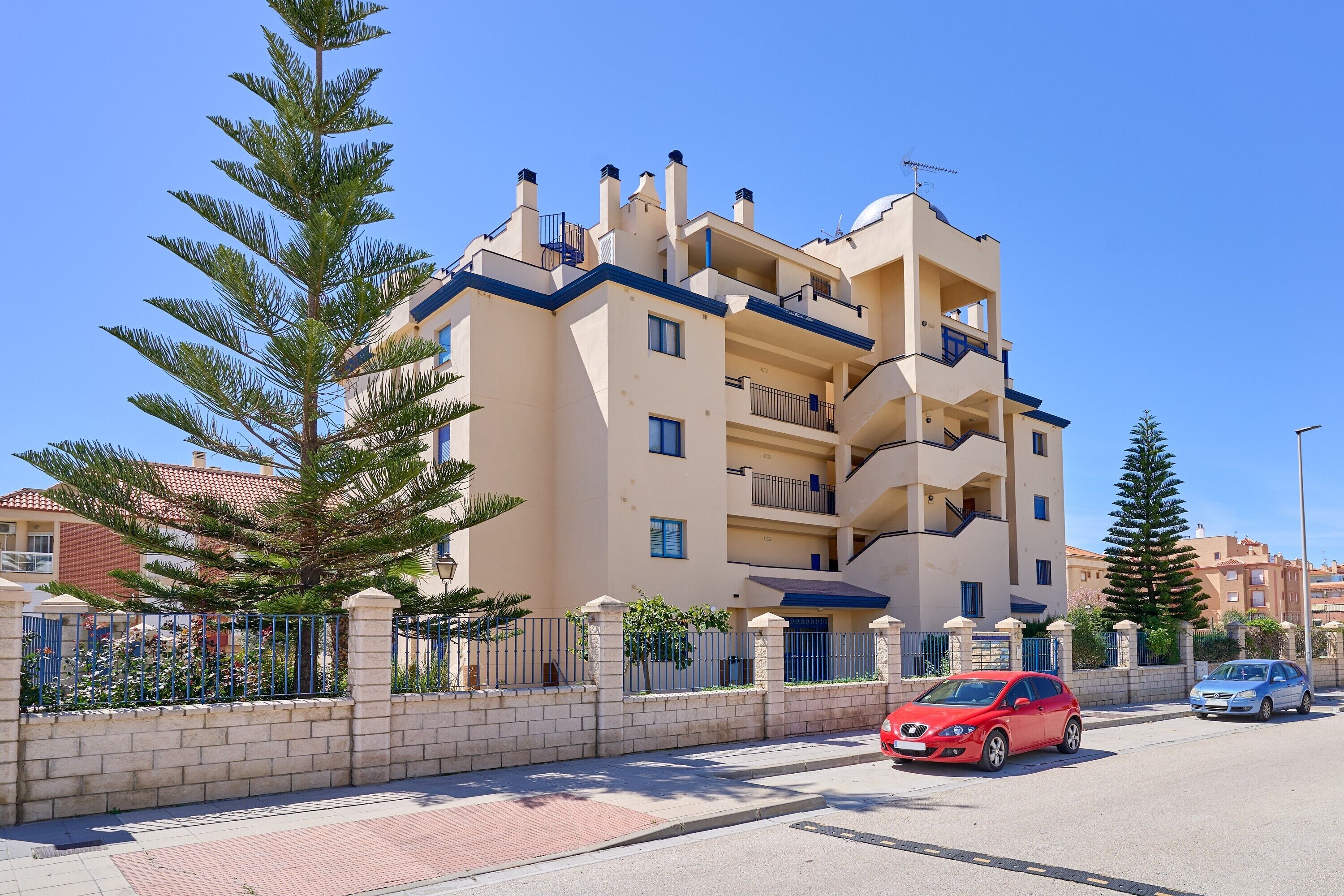 Un edificio residencial de varios pisos con balcones, una acera pavimentada y una calle con coches estacionados.