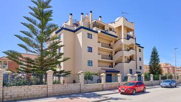 Un edificio residencial de varios pisos con balcones, una acera pavimentada y una calle con coches estacionados.