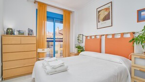 A hotel room with a bed, a wooden dresser, a window with curtains, a framed picture on the wall, and a potted plant. - Apartment 'Argia' with Sea View, Wi-Fi, and Air Conditioning (Algarrobo)