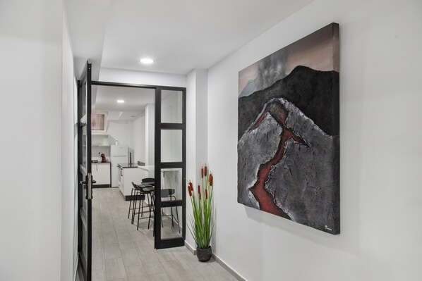 A hallway with a modern kitchen in the background, a potted plant, and a large abstract painting on the wall.