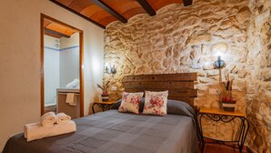 A bedroom with a stone wall, a wooden headboard, a bed with a grey bedspread, two bedside tables with lamps, and a bathroom visible through an open door. - Country House 'Virginia' with Mountain View and Balcony (Unknown)