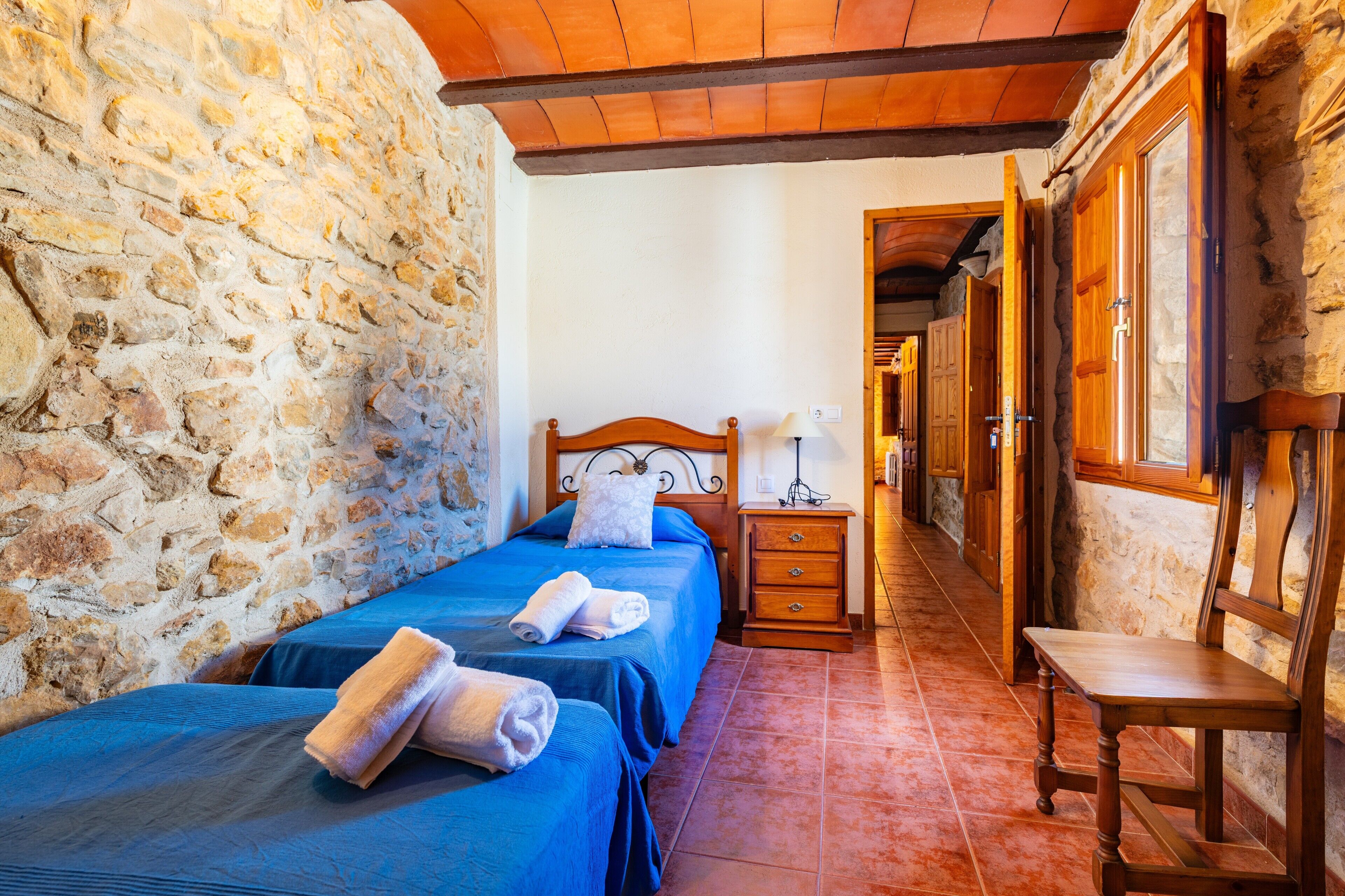 Una habitación con paredes de piedra, techo de madera, una cama con ropa de cama azul, una mesita de noche de madera y un banco.
