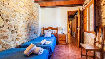 Una habitación con paredes de piedra, techo de madera, una cama con ropa de cama azul, una mesita de noche de madera y un banco.