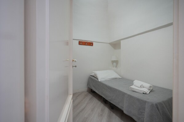 A narrow room with a single bed, a small table, and a wall-mounted light fixture.