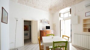 Een eetgedeelte met een witte tafel en stoelen, een houten kast, een vloerlamp en een keuken op de achtergrond.