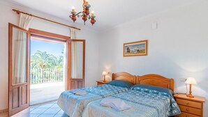 Un dormitorio con una cama doble, una mesita de noche de madera con una lámpara, vistas a palmeras a través de una puerta abierta y un cuadro en la pared.