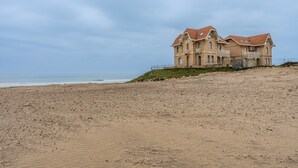 Een groot, twee verdiepingen tellend gebouw met een rood pannendak, gelegen op een zandstrand met de oceaan op de achtergrond.