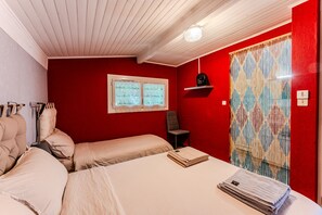 Ein Zimmer mit zwei Betten, einem Fenster, einem Sessel, einem Regal mit einer Dekoration und einem gemusterten Vorhang.