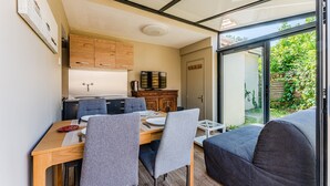 Een compacte keuken met moderne apparaten, een eetgedeelte met grijs beklede stoelen en uitzicht op groen door een glazen deur.