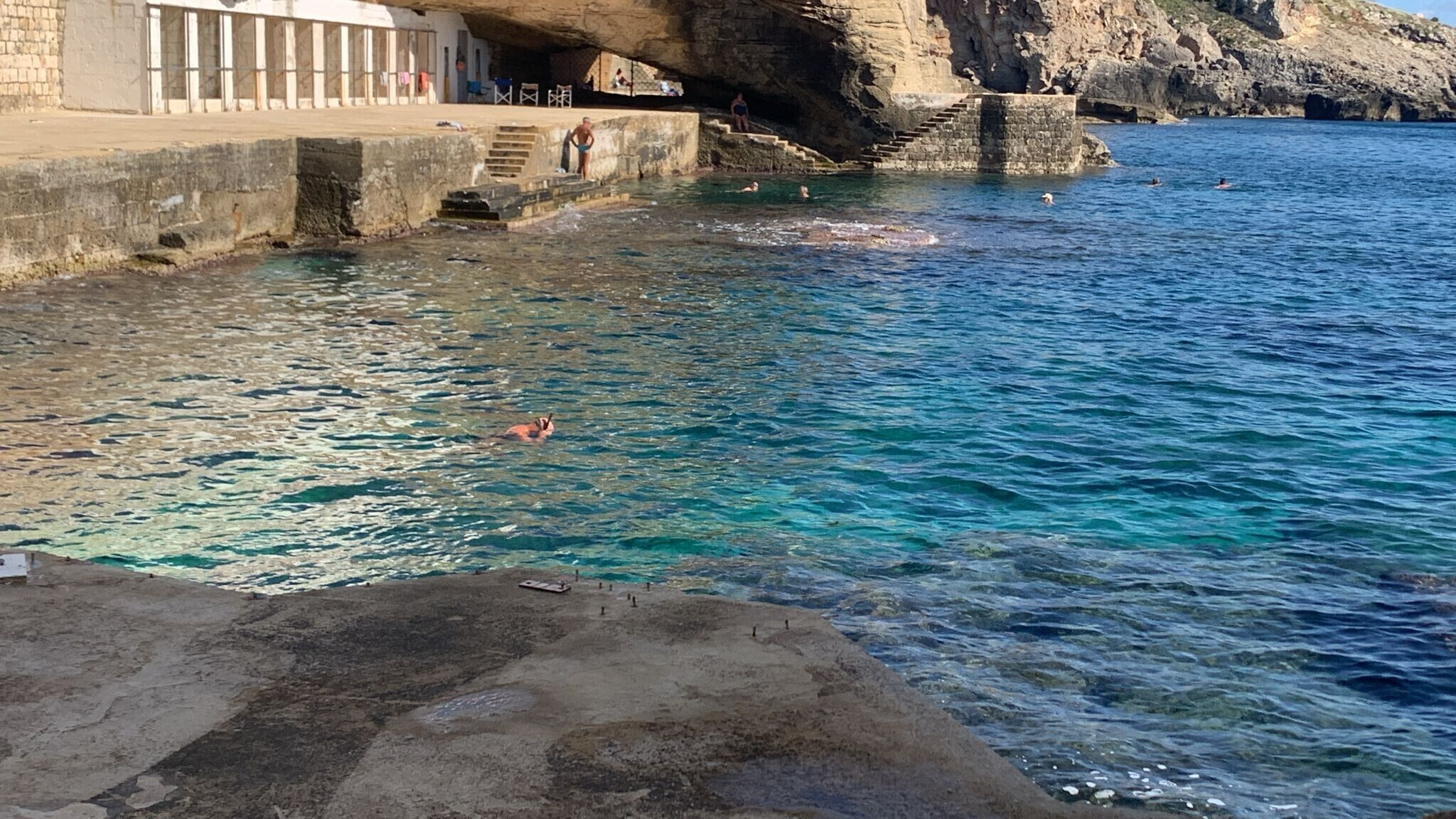 Una zona costera con piscina, escalones que bajan al agua y un acantilado rocoso.