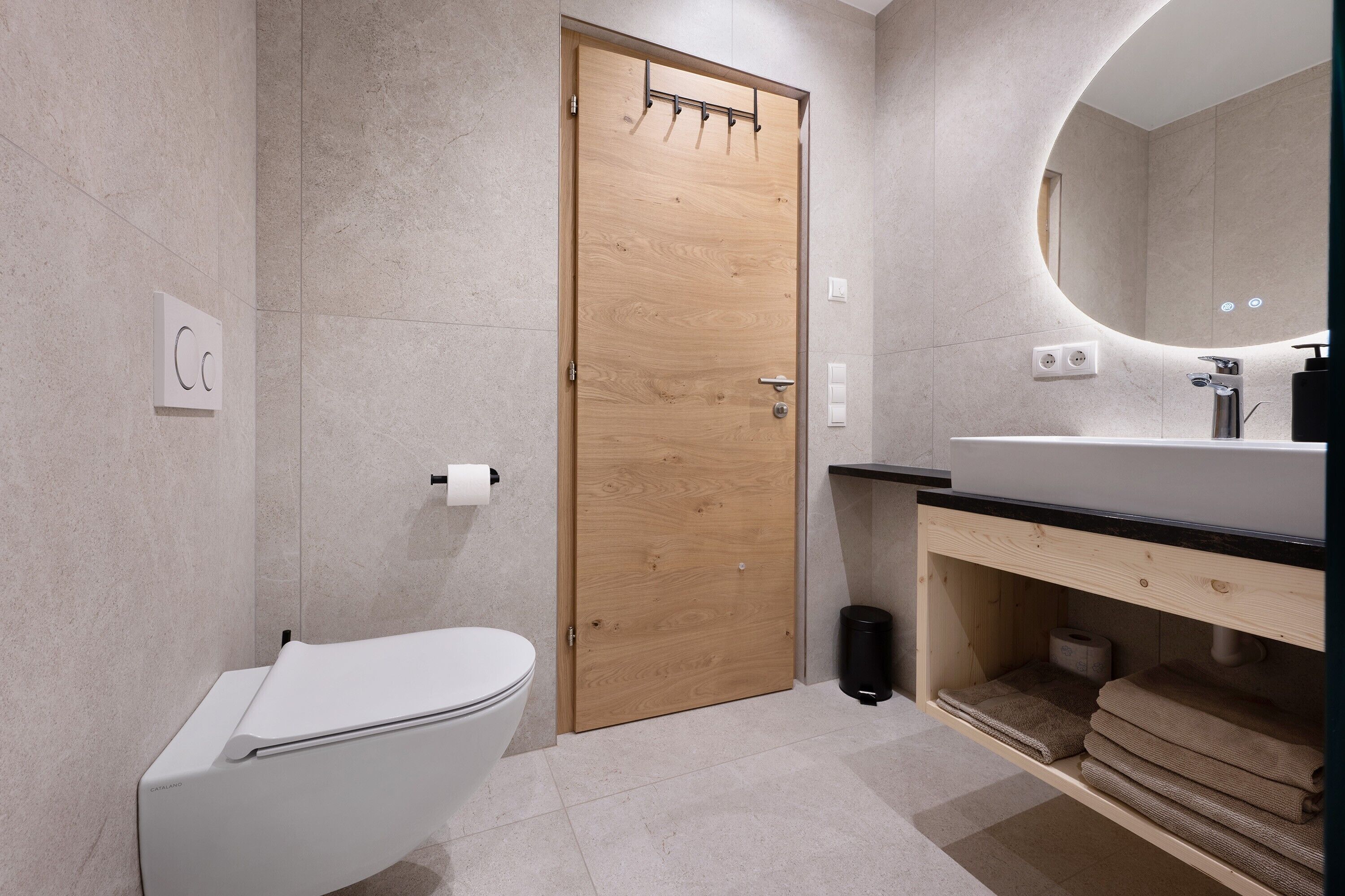 Et moderne badeværelse med dør i træ, et hvidt toilet, en vask med sort bordplade og et spejl.