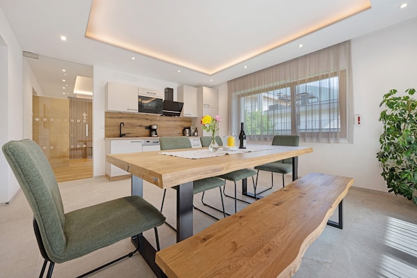 A modern dining area with a wooden table, green upholstered chairs, and a large window with sheer curtains.