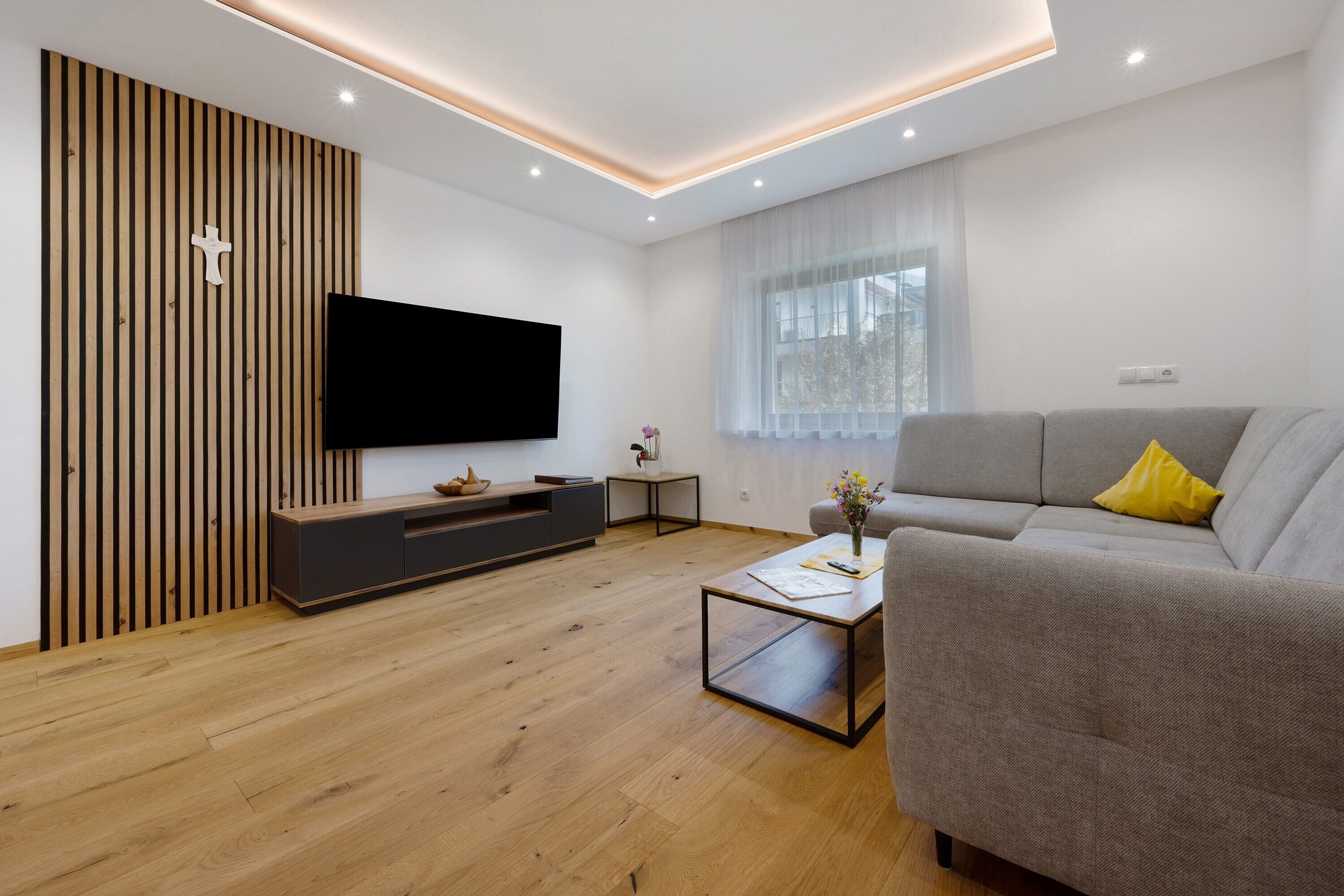 A modern living room with a flat-screen TV, grey sofa, wooden floor, and a window with sheer curtains.