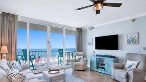 Ein geräumiges Wohnzimmer mit einem großen Schiebeglasfenster, das auf einen Balkon mit Blick aufs Meer führt. Es gibt einen Deckenventilator, einen flachbildfernseher an der Wand und ein helles Sofa mit gestreiften Kissen.