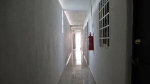 Un pasillo largo y estrecho con paredes blancas y suelo de baldosas. Un extintor rojo está fijado en la pared a la derecha.