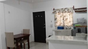 Una pequeña kitchenette con microondas, un lavamanos y un ventanal con cortinas. Hay una mesa de comedor con sillas y una puerta negra.