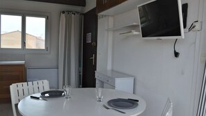 A small, clean room with a white table set for two, a TV mounted on the wall, and a window with curtains.