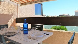 A table set for two with a blue bottle, glasses, and cutlery on a balcony overlooking a tennis court.