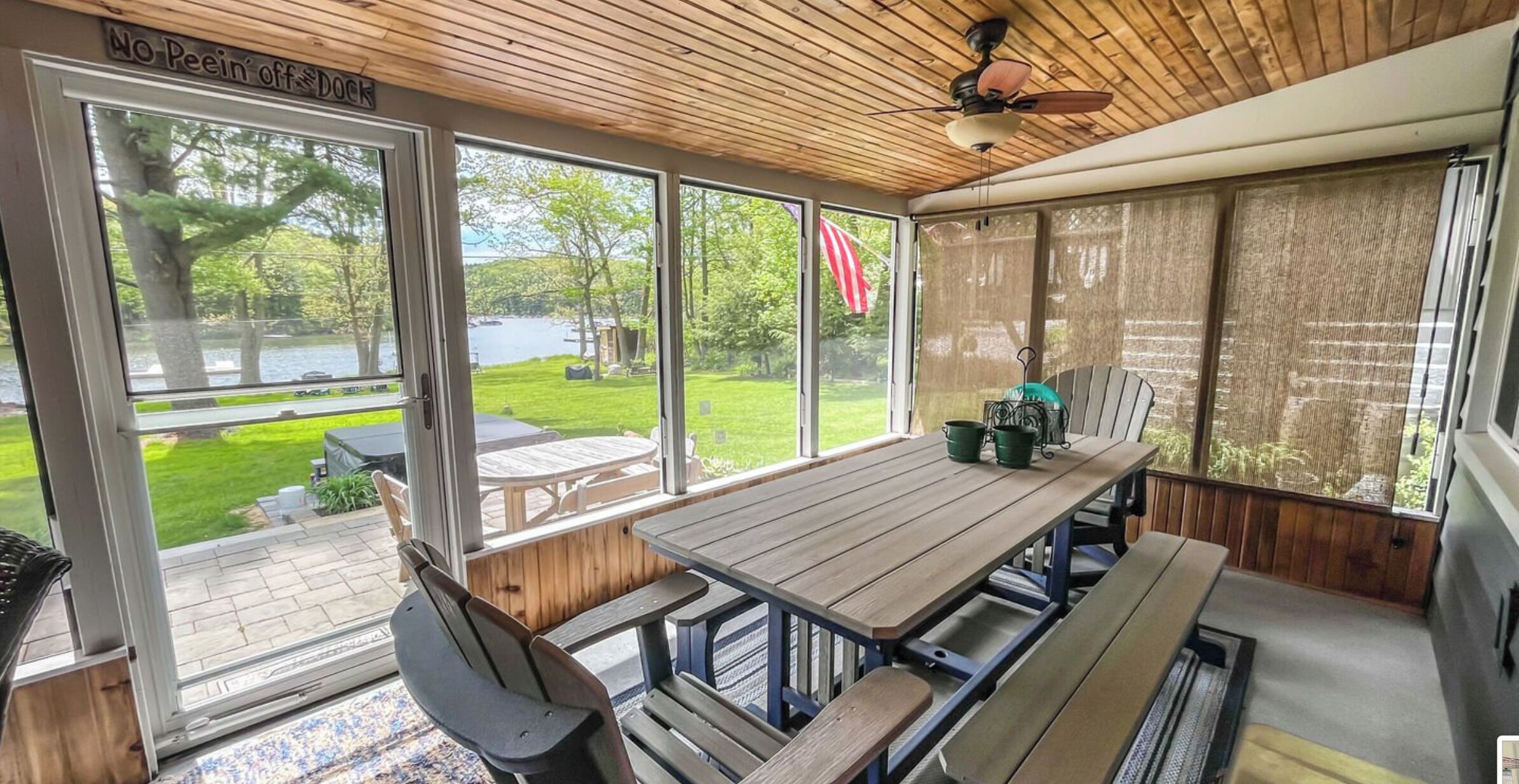 Un patio couvert avec une table et des bancs en bois, de grandes fenêtres offrant une vue sur un lac et la verdure, et un panneau indiquant : « Il est interdit de faire ses besoins sur le quai ».
