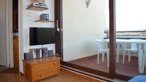 A living room with a TV, wooden cabinet, and a balcony with plastic chairs. - Appart 2 pièces 4 couchages PORT LEUCATE NATURISME (PORT LEUCATE NATURISME)