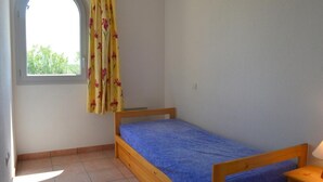 A small room with a single bed, a window with a curtain, and a lamp on a bedside table. - Mais 3 pièces 6 couchages PORT LEUCATE NATURISME (PORT LEUCATE NATURISME)