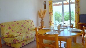 A dining area with a yellow floral sofa, wooden dining table, and chairs. There is a vase with dried plants on the table, and a clock on the wall. - Mais 3 pièces 6 couchages PORT LEUCATE NATURISME (PORT LEUCATE NATURISME)