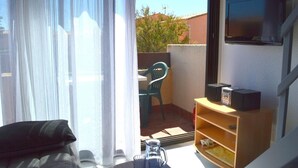 A room with a TV mounted on the wall, a small wooden shelf with electronic devices, a chair, and a table outside a sliding glass door. - Appart Studio / mezz 2 couchages PORT LEUCATE NATURISME (PORT LEUCATE NATURISME)
