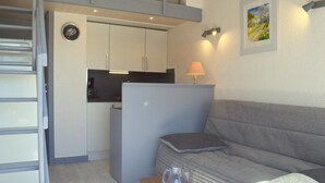 A compact living space with a kitchenette, a sofa, a small table, and a lamp. - Appart Studio / mezz 2 couchages PORT LEUCATE NATURISME (PORT LEUCATE NATURISME)