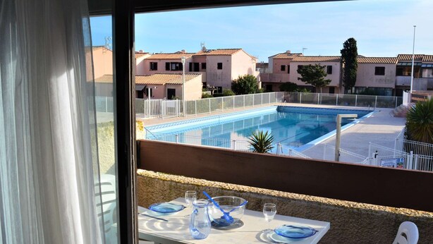 A balcony with a table set for two, overlooking a swimming pool and residential buildings. - Appart Studio / mezz 2 couchages PORT LEUCATE NATURISME (PORT LEUCATE NATURISME)