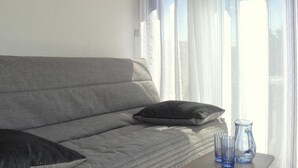 A grey sofa with a cushion, a small table with two blue glass vases, and sheer curtains on a window. - Appart Studio / mezz 2 couchages PORT LEUCATE NATURISME (PORT LEUCATE NATURISME)