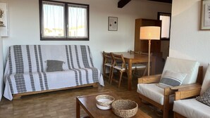 Ein gemütliches Wohnzimmer mit einem gemusterten Sofa, einem Holzesstisch und Stühlen. Auf dem Tisch steht eine Lampe und es gibt ein Fenster mit Vorhängen.