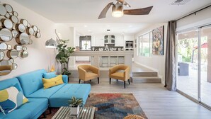 Una sala moderna con un sofá azul, cojines amarillos y un tapete estampado. Hay una zona de cocina con armarios blancos y un rincón comedor con dos sillas amarillas.