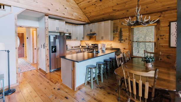 A wooden cabin interior with a kitchen, dining area, and a large window.