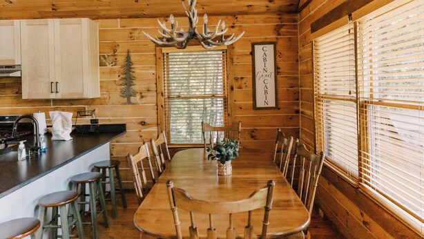 A wooden cabin interior with a dining table, chairs, a chandelier, and a kitchen area.