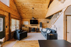 A cozy cabin interior with a wooden ceiling, a sofa, a dining table, a fireplace, and a staircase.