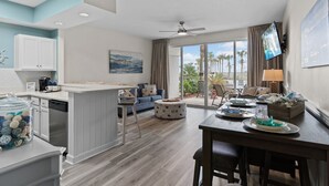 Een moderne keuken met witte kasten, een eethoek met een houten tafel en een zithoek met een blauwe bank en een groot raam met uitzicht op palmbomen.